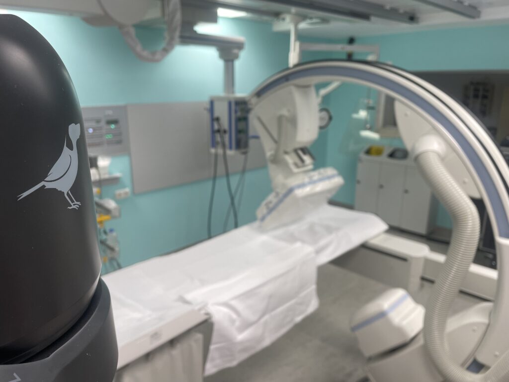 Black PTZ camera in the foreground, a medical procedure room with C-arm and patient table behind it