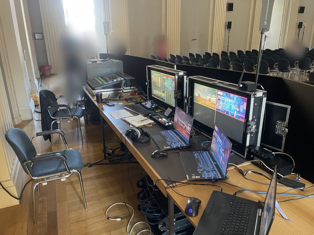 Event control desk with monitors in flight cases, laptops and control panels, a hall with rows of chairs behind it